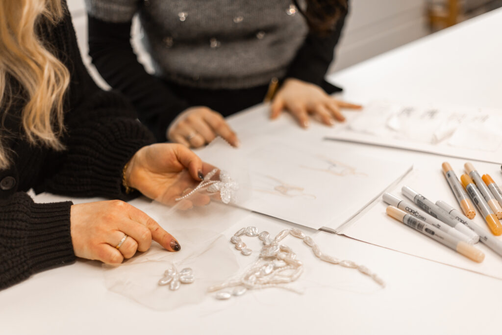 Zwei Personen schauen über Designentwürfe für Brautkleider, mit Copics und Papier auf dem Tisch