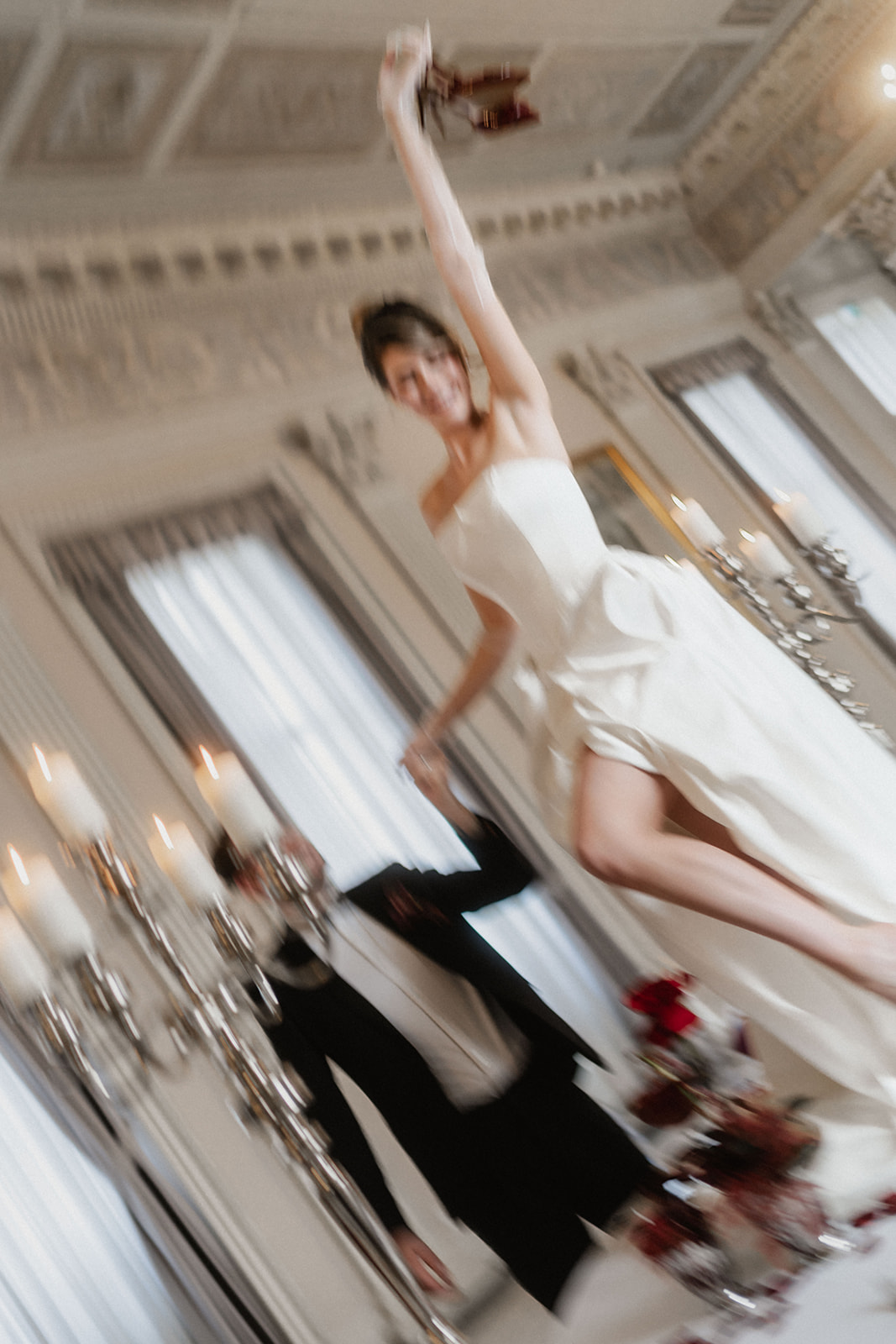 Verwischtes Bild eines Brautpaares. Er im Anzug, Sie in einem Kleid von Lisa Dörr Couture mit Schlitz. Sie steht auf einem Tisch und wirft dir Hand in die Höhe. Vintage inspiriert.
